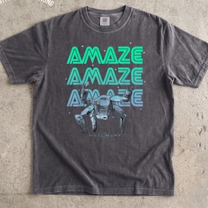 May include: A charcoal gray short-sleeve t-shirt featuring the text AMAZE in a repeating teal and light blue gradient font. Below the text is a graphic of a metallic crab-like creature and the small text HAIL MARY. SWM CUSTOM CLOTHING is printed.