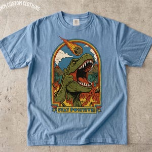 May include: A light blue t-shirt featuring a graphic of a green T-Rex giving a thumbs up with an open mouth. Above the dinosaur is a flaming meteor, and below is the text "STAY POSITIVE!" framed by an arch with palm trees and a volcano.