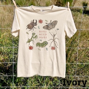 May include: An ivory short-sleeve t-shirt featuring a graphic print of various insects including a beetle, moth, praying mantis, caterpillar, and stick insect. The text "BUGS NEED HUGS" is printed in red in the center, surrounded by small red stars.