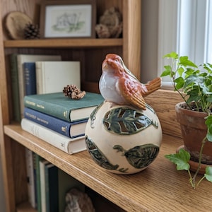 May include: A ceramic bird figurine with a reddish-brown head and wings perched atop a spherical base. The base is cream-colored with cut-out leaf patterns in dark green, featuring a glossy finish on the bird and a matte finish on the sphere.