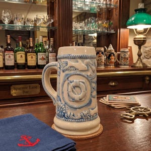 May include: A cream-colored beer stein with blue nautical relief designs, including a life preserver and rope, sits on a wooden surface. The mug has a large handle and is placed on a cork coaster. A blue napkin with a red anchor is in the foreground.