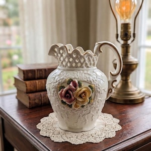 May include: A white ceramic pitcher with a decorative floral design and a handle. The pitcher features a cluster of pink and yellow roses. It sits on a lace doily on a wooden table. A vintage lamp is in the background.