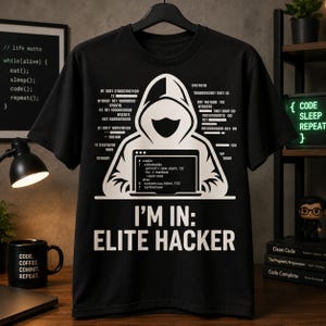 Op de afbeelding: Zwart t-shirt met een witte afbeelding van een figuur met een capuchon die op een laptop werkt. De tekst "I'M IN: ELITE HACKER" staat eronder. Binaire code en programmeerfragmenten zijn ook op het shirt gedrukt.