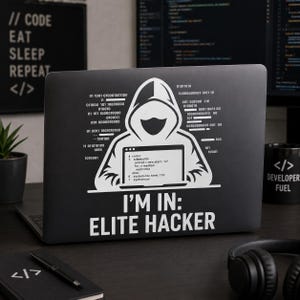 Op de afbeelding: Zwarte laptop met een witte afbeelding van een figuur met capuchon en de tekst "I'M IN: ELITE HACKER." Binaire code en programmeertermen zijn ook zichtbaar. Een mok met "DEVELOPER FUEL" is zichtbaar.