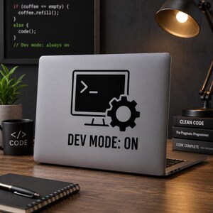 Op de afbeelding: Een laptop met een zwarte "DEV MODE: ON" sticker met een computermonitorpictogram en een tandwiel. Links staat een zwarte mok met "</> CODE". Boeken en een bureaulamp op de achtergrond.