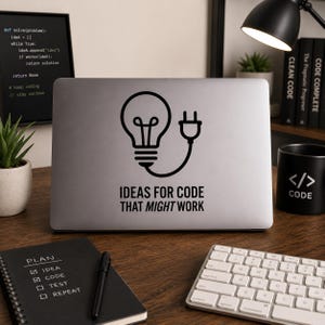 Op de afbeelding: Een laptop met een zwarte afbeelding van een gloeilamp en een stekker, met de tekst "IDEAS FOR CODE THAT MIGHT WORK". Een zwarte mok met codesymbolen, een toetsenbord en een notitieboek zijn ook zichtbaar.