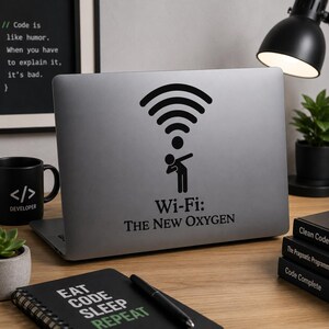 Op de afbeelding: Een zilveren laptop met een zwarte Wi-Fi-signaalsticker en de tekst "Wi-Fi: The New Oxygen". Een zwarte mok met de tekst "</> DEVELOPER" en een notitieboek met "EAT CODE SLEEP REPEAT" zijn ook zichtbaar.