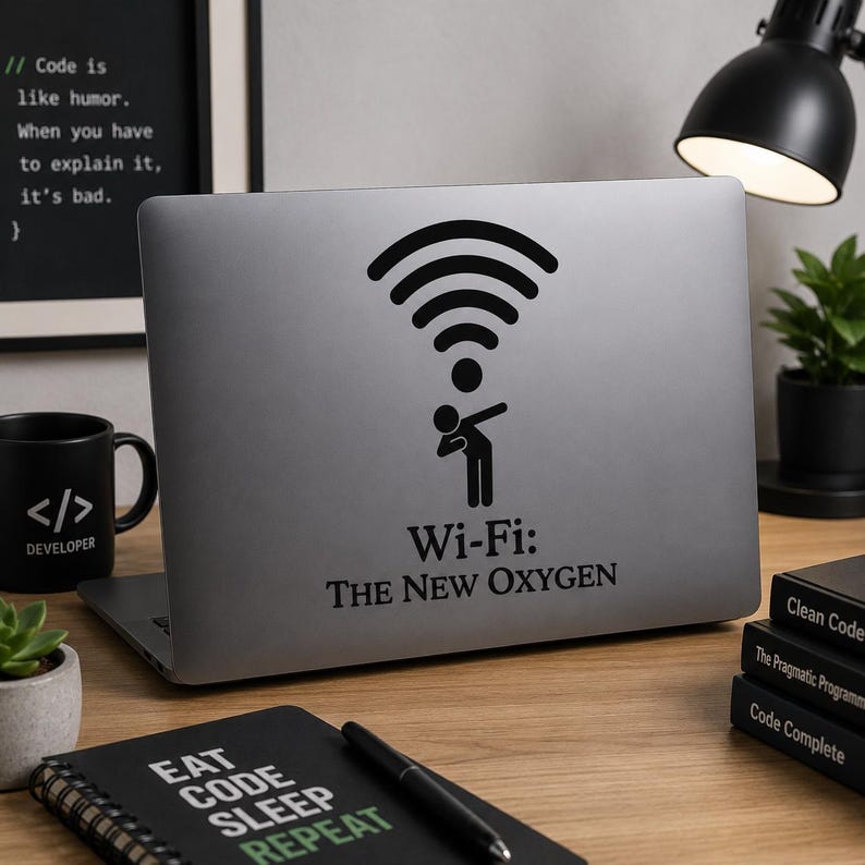 Op de afbeelding: Een zilveren laptop met een zwarte Wi-Fi-signaalsticker en de tekst "Wi-Fi: The New Oxygen". Een zwarte mok met de tekst "</> DEVELOPER" en een notitieboek met "EAT CODE SLEEP REPEAT" zijn ook zichtbaar.