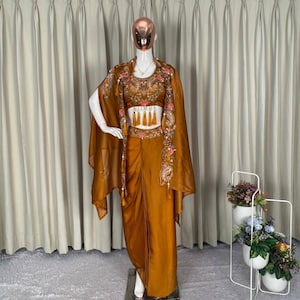 May include: A mustard-coloured outfit is displayed on a mannequin. The ensemble features a crop top with floral embroidery, a matching cape, and draped trousers. The cape and top have detailed embellishments with tassels. The backdrop is a neutral curtain.