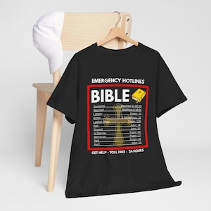 May include: Black t-shirt with the text "EMERGENCY HOTLINES BIBLE" in white. The shirt features a cross graphic and a list of Bible verses for various emotional states. A white baseball cap hangs on a wooden chair.
