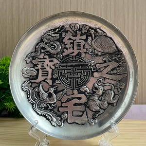 Puede incluir: Un plato circular de metal plateado con relieves esculpidos de un dragón, una tortuga, un fénix y un tigre que rodean un símbolo geométrico central. Los caracteres chinos 鎮宅之寶 se muestran de forma prominente en el centro.