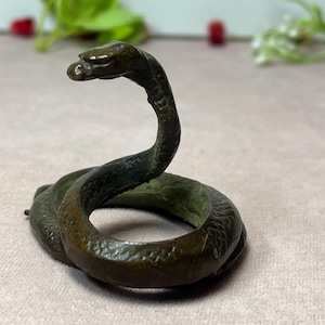 Op de afbeelding: Een klein, donker bronskleurig metalen beeldje van een opgerolde slang. De slang is afgebeeld in een rechtopstaande, alerte houding met de kop omhoog en gebogen, met gedetailleerde schubstructuren over het hele lichaam en een donkere afwerking.