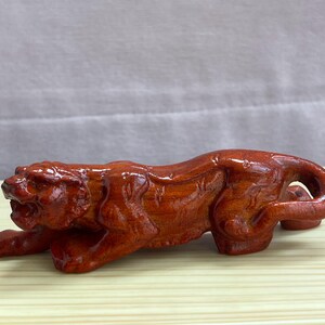 Può includere: Una scultura in legno levigato color bruno-rossastro raffigurante una tigre accovacciata con la bocca aperta, che mostra tratti del volto dettagliati e un corpo muscoloso. La statuetta ha una finitura liscia e lucida che esalta le venature.