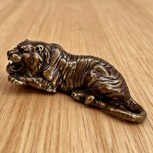 Op de afbeelding: Een bronzen tijgerbeeldje in een ontspannen pose, met gedetailleerde vachttextuur en een open mond. De sculptuur ligt, met zijn poten en staart zichtbaar. Het beeldje is een decoratief item.