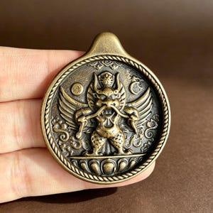 Peut inclure: Un pendentif circulaire en métal de couleur bronze présentant un relief d'une créature ailée avec des cornes et une longue moustache. Le design comprend une bordure en corde, une base en lotus et des symboles célestes comme un soleil et une lune.