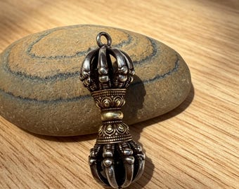 May include: A silver-toned pendant with a crown-like top and a detailed, ornate design. The pendant has a gold-colored band in the middle and a claw-like bottom. The pendant is resting on a smooth, gray stone, with a wooden surface in the background.
