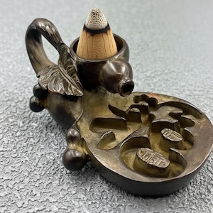 Pode incluir: Um queimador de incenso em tom bronze com um design semelhante a um cisne, com um incenso em forma de cone. O queimador tem detalhes intrincados, incluindo uma folha e uma base decorativa com padrões esculpidos. O incenso é castanho claro com uma ponta escura.
