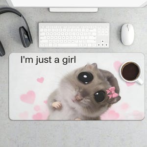May include: A rectangular mousepad with the text "I'm just a girl" and a cartoon illustration of a gray hamster with large black eyes and a pink bow. Pink hearts are scattered across the white background. A cup of coffee is on the right.