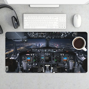 May include: A rectangular desk mat featuring a detailed image of an airplane cockpit at night, with a cityscape visible through the windshield. A white coffee mug sits on the right side of the mat.