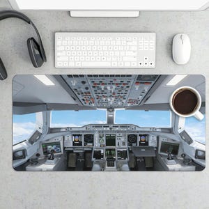 May include: A desk mat featuring a detailed image of an airplane cockpit, complete with instrument panels, screens, and controls. A cup of coffee sits on the mat. A keyboard, mouse, and headphones are also visible.