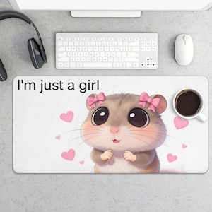 May include: White rectangular mousepad featuring a cartoon hamster with pink bows and hearts, and the text "I'm just a girl." A cup of coffee sits on the right. A keyboard, mouse, and headphones are visible.