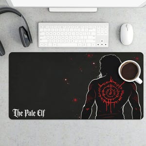 May include: Black mousepad featuring a stylized illustration of a person with a red tattoo design on their back. The text "The Pale Elf" is printed in white. A cup of coffee sits on the right side.
