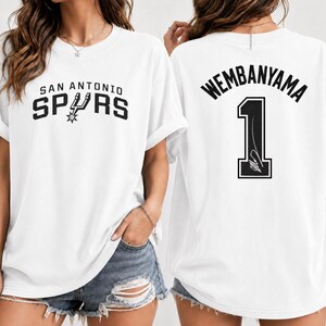 Pu&ograve; includere: Una t-shirt bianca a maniche corte con il logo e la scritta San Antonio Spurs sul davanti. Il retro della maglietta mostra il nome Wembanyama in nero sopra il numero 1, che include un piccolo grafico di una firma all'interno della cifra.