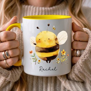 May include: White ceramic mug with a yellow interior and handle, featuring a cartoon bee illustration. The bee is yellow and black with white wings, surrounded by flowers and the name "Rachel".