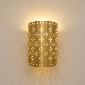 May include: A semi-cylindrical wall sconce made of brushed gold metal featuring a repeating pattern of small, diamond-shaped cutouts. The fixture emits a warm, soft glow from the top and bottom, casting light through the perforations.