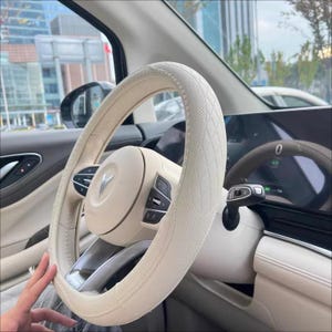 May include: Cream-colored car steering wheel with a diamond-stitched cover. The steering wheel has integrated controls and a central emblem. The interior of the car is visible, with a digital display and other controls.