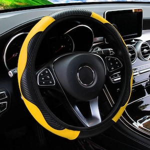 May include: A car steering wheel with a yellow and black cover. The cover features a carbon fibre texture on the black sections and a smooth yellow finish. The steering wheel is black with various control buttons.
