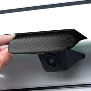 May include: A black carbon fiber textured car accessory, possibly a camera cover or trim piece. The accessory is held up against a white car surface, with a camera visible below.