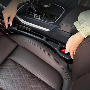 May include: A black car seat organizer with a red release button is installed between the front seats. The car seat is brown leather with a diamond pattern. A hand is shown inserting the organizer.