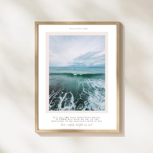May include: A framed print displaying a tranquil ocean view with turquoise water and a cloudy sky. The print includes the text "Psalm 91:16 NIV" at the top and a Bible verse at the bottom. The frame is a light wood colour.