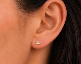 May include: A close-up of a person's ear with a small gold stud earring. The earring features three small, round, clear gemstones arranged in a triangular shape. The earring is set close to the earlobe.