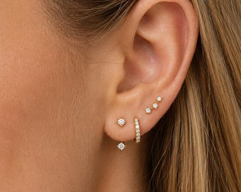 May include: Close-up of a person's ear adorned with multiple gold earrings. The earrings include small studs, a hoop, and a graduated line of three small studs, all featuring sparkling clear stones.