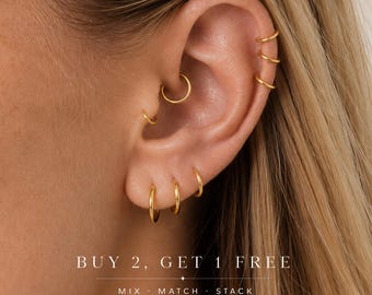 May include: A set of gold hoop earrings worn in multiple ear piercings, including the lobe, tragus, and helix. The text on the image reads: BUY 2, GET 1 FREE. MIX . MATCH . STACK. The earrings are thin, circular, and polished gold metal.