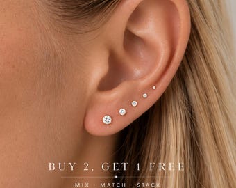 May include: A row of five graduated round clear stud earrings set in gold metal, worn along the earlobe and cartilage. The text reads: BUY 2, GET 1 FREE. MIX · MATCH · STACK.