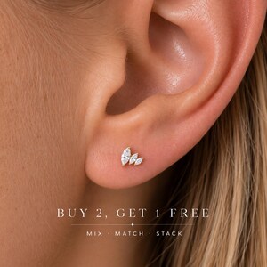 May include: A gold stud earring featuring three marquise-cut clear stones arranged in a fan shape. The text BUY 2, GET 1 FREE and MIX · MATCH · STACK is printed below the earring. The earring is positioned on an earlobe.