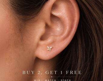 May include: A small gold butterfly stud earring featuring four clear, sparkling stones arranged in a wing pattern, worn in the earlobe. The image includes the text: BUY 2, GET 1 FREE, MIX · MATCH · STACK.