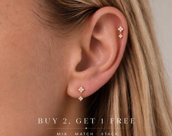 May include: Two gold-toned earrings featuring a small four-stone cluster design with clear gems. One earring is placed in the earlobe and the other in the upper cartilage. The text reads: BUY 2, GET 1 FREE. MIX · MATCH · STACK.