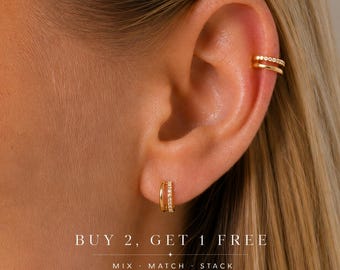 May include: A gold ear cuff with a row of small clear stones and a matching gold hoop earring with a row of clear stones on the earlobe. The text reads: BUY 2, GET 1 FREE. MIX · MATCH · STACK.
