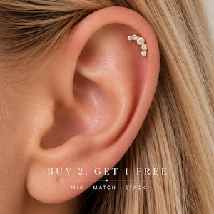 May include: A gold cartilage earring featuring a curved row of five round clear stones of varying sizes. The jewelry is worn in the upper ear. Text on the image reads: BUY 2, GET 1 FREE. MIX MATCH STACK.