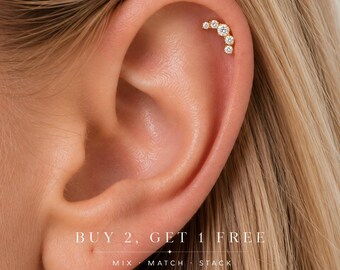 May include: A gold cartilage earring featuring a curved row of five round clear stones of varying sizes. The jewelry is worn in the upper ear. Text on the image reads: BUY 2, GET 1 FREE. MIX MATCH STACK.