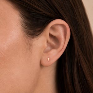 May include: A small, gold-colored cross stud earring is positioned in the lower lobe of an ear. The earring features a simple, minimalist cross shape with a polished finish, centered on the earlobe against the skin.