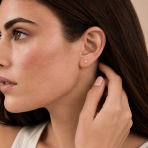 May include: A small, gold-colored stud earring in the shape of a cross. The earring is positioned on the earlobe, featuring a simple, minimalist design with a polished metallic finish that reflects light, creating a subtle and understated appearance.