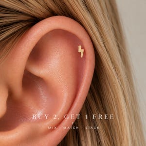 May include: A small gold-colored lightning bolt stud earring worn in the upper cartilage of an ear. The image includes the text: BUY 2, GET 1 FREE, MIX · MATCH · STACK. The earring features a simple, flat, geometric lightning bolt shape.