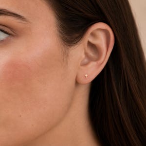 May include: A small, gold-colored stud earring in the shape of a cross, positioned in the lower lobe of an ear. The earring features a simple, minimalist design with a polished finish, reflecting light on its vertical and horizontal bars.