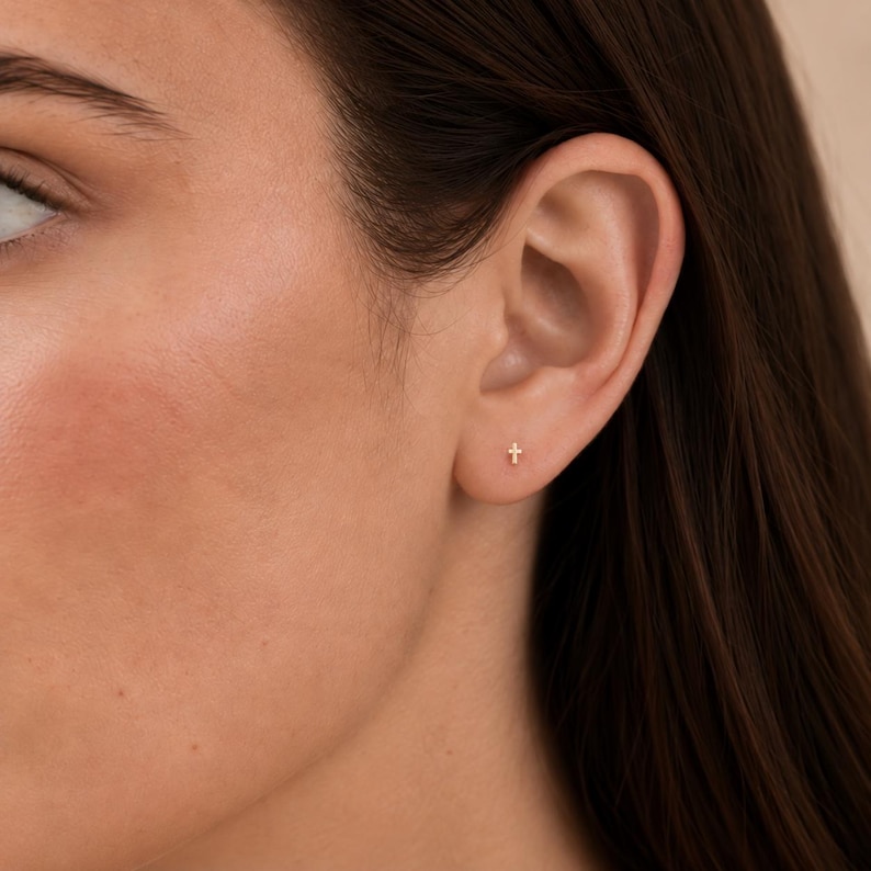 May include: A small, gold-colored stud earring in the shape of a cross, positioned in the lower lobe of an ear. The earring features a simple, minimalist design with a polished finish, reflecting light on its vertical and horizontal bars.