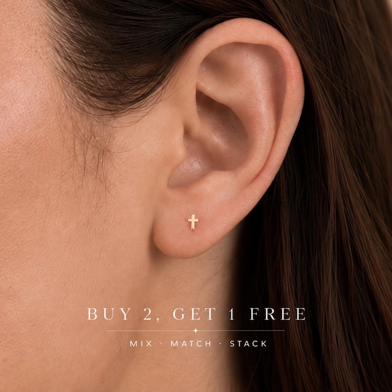 May include: A small gold-toned cross stud earring worn in the earlobe. The image includes the text BUY 2, GET 1 FREE, MIX &middot; MATCH &middot; STACK at the bottom, featuring a minimalist cross design with a polished finish and a simple post backing.